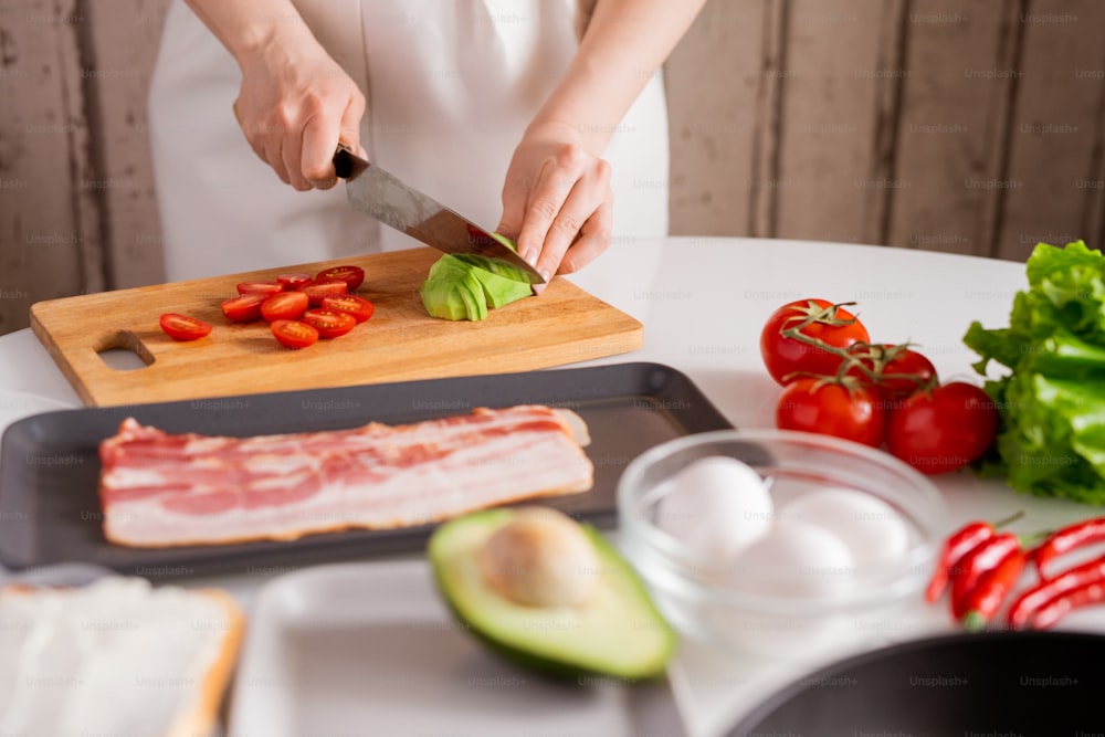 Hands of young woman standing by table in the kitchen and cutting fresh avocado and tomatoes on chopping board while cooking salad