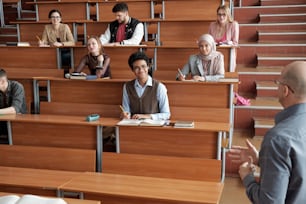 Felices estudiantes universitarios interculturales sentados junto a largos escritorios de madera en la sala de conferencias, tomando notas y escuchando al profesor en la clase