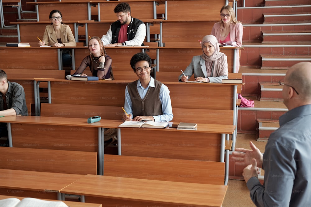 Happy intercultural university students sitting by long wooden desks in lecture hall, making notes and listening to professor at lesson