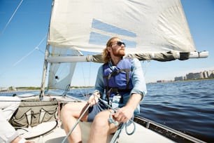 Jeune homme hipster sérieux et pensif avec une barbe tenant une corde tout en conduisant un voilier et en profitant du paysage autour