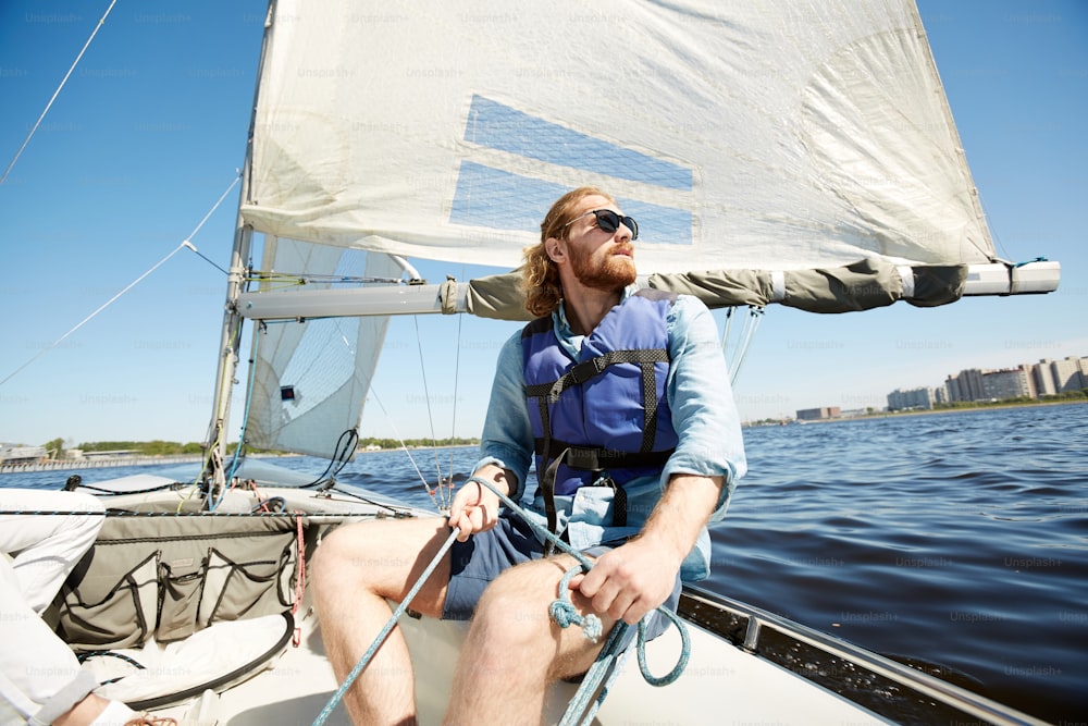 Serio giovane hipster pensieroso con la corda che tiene la barba mentre si opera la barca a vela e si gode il paesaggio intorno