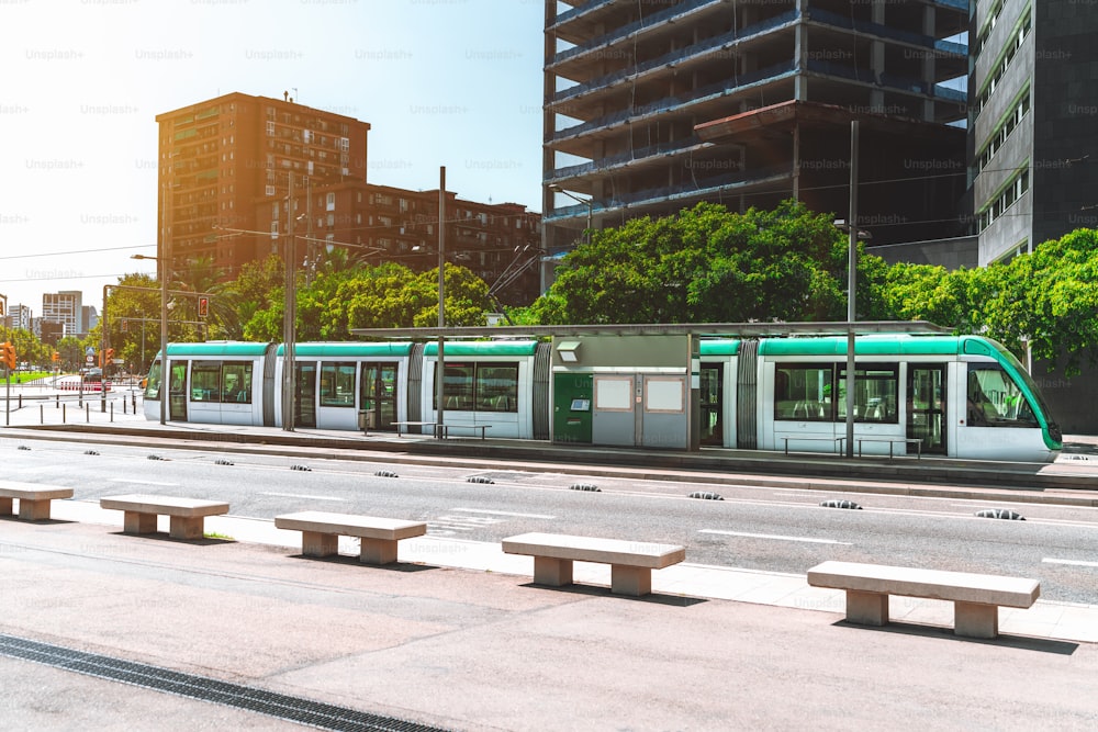 Wide-angle view of a modern city tram in urban settings; green and white city streetcar with the row of concrete benches in front; contemporary tramcar on the station with the road in the foreground