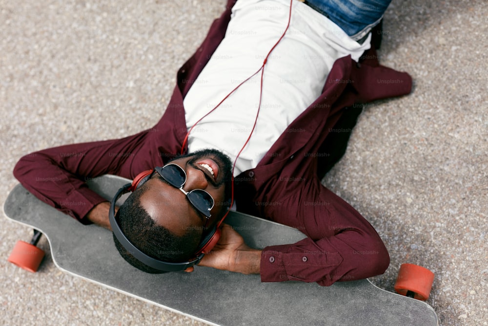 Mode. Homme noir écoutant de la musique dans des écouteurs dans la rue, allongé sur du longboard. Haute résolution