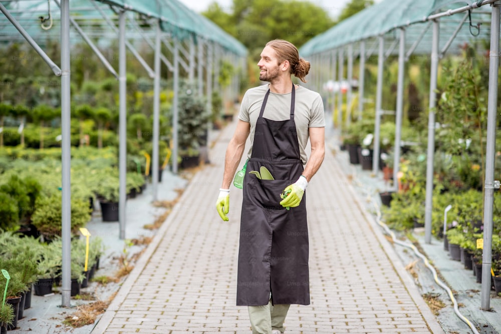 Bel lavoratore in uniforme che cammina alla serra con piante verdi in vendita