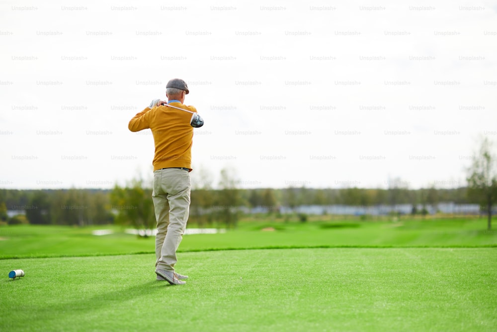 Vista posteriore dell'uomo anziano in berretto, pullover giallo e pantaloni beige palla colpita con mazza da golf sul campo verde