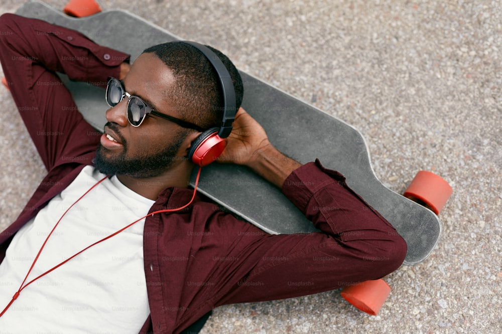 Moda. Uomo nero che ascolta musica in cuffia sulla strada, sdraiato sul longboard. Alta risoluzione