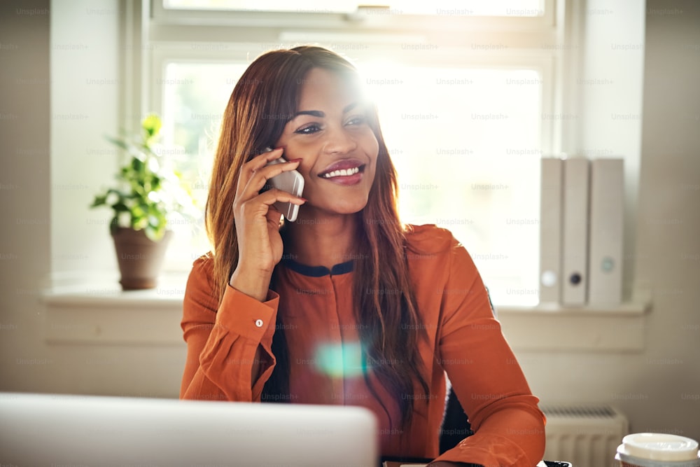 Smiling young African female entrepreneur sitting at a table in her home office working on a laptop and talking on a cellphone