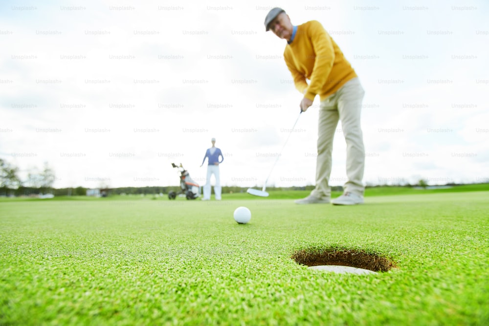 Mature man in casualwear standing on green field and going to hit golf ball into hole