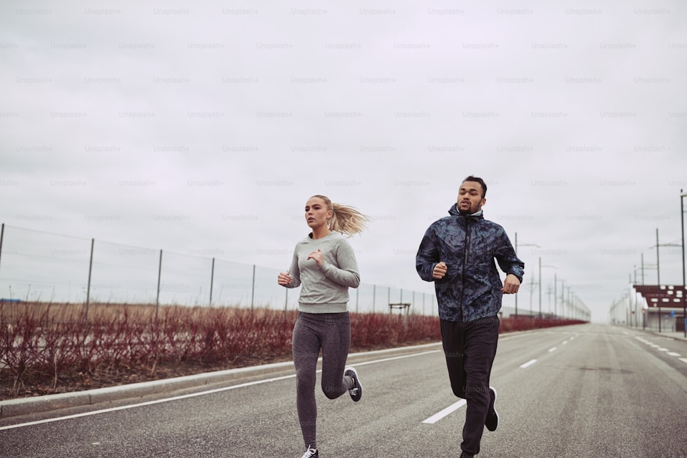 Jovem casal diverso em roupas esportivas correndo juntos ao longo de uma estrada no país em um dia nublado