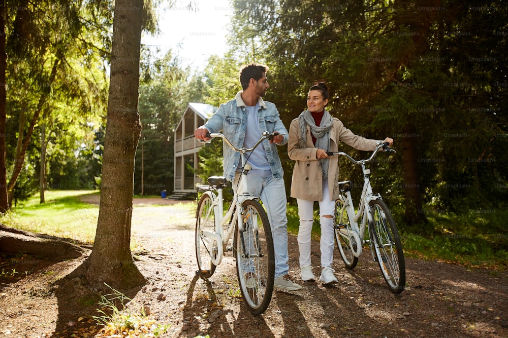 Smiling loving young couple in casual clothing walking with bicycles in autumn forest and looking at each other enjoying sunny day