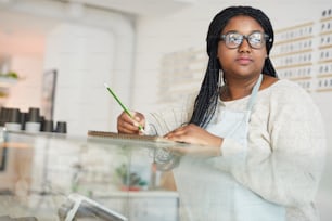 Young mixed-race woman standing by display with pastry and making working notes in notepad