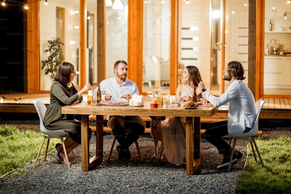 야외에서 저녁 시간에 조명으로 장식된 현대적인 집의 뒷마당에 있는 테이블에 앉아 저녁을 먹는 친구들