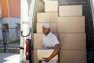 Courier Delivery Service. Portrait Of Black Man Delivering Package Near Car With Boxes Outdoors. High Resolution