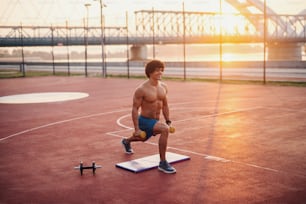 早朝に運動する若い決定的なフィットマン。ウェイトを手にした屋外のトレーニングフィールドでのトレーニング。
