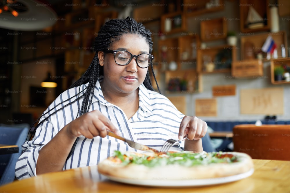 Hungry mixed-race girl with African braids sitting by table and slicing pizza before eating