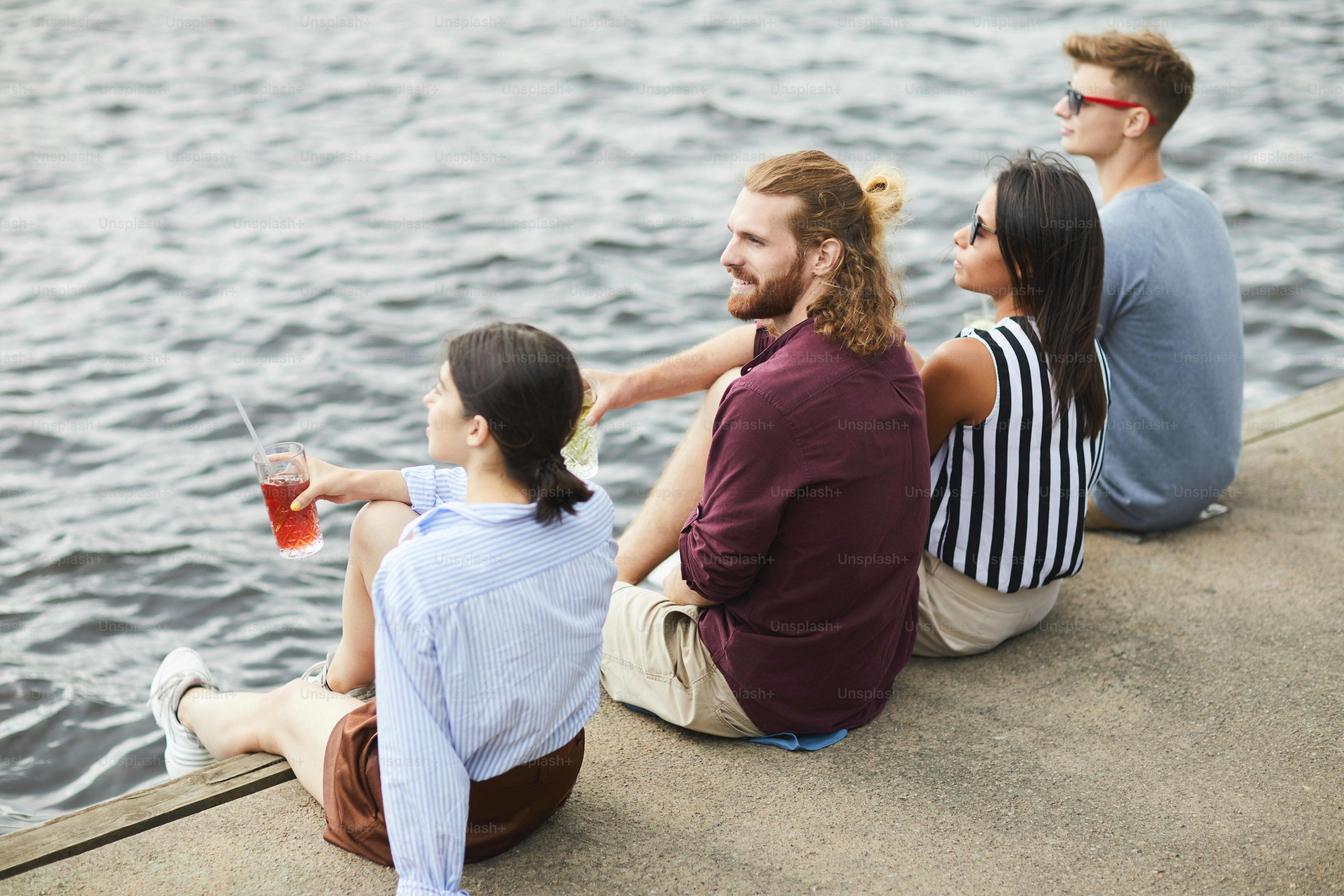 Back view of two casual couples sitting on wooden pontoon by waterside and having drinks