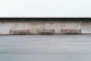 Scatto minimalista grandangolare di tre panchine di legno con gambe metalliche in piedi di fronte a un muro di cemento grunge con piccoli fori, un'enorme area vuota dell'asfalto con una singola striscia in primo piano