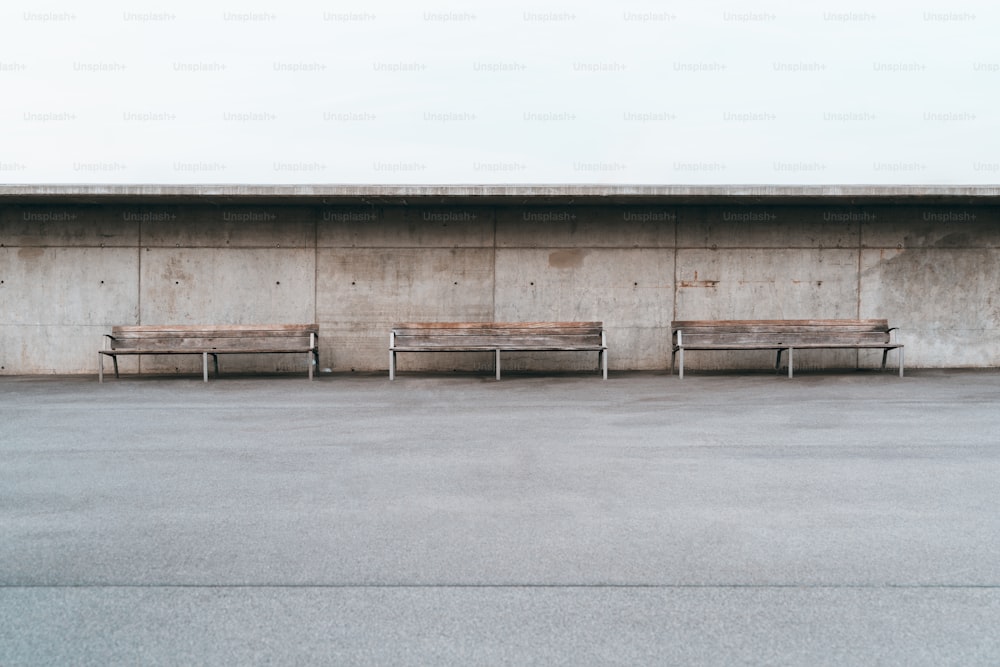 Wide-angle minimalist shot of three wooden benches with metal legs standing in front of a grunge concrete wall with small holes, a huge empty area of the asphalt with a single stripe in the foreground