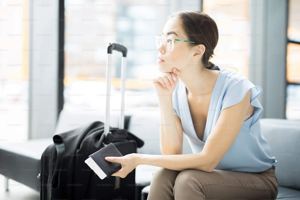 Jeune voyageuse d’affaires pensive avec passeport et billets assise dans le salon de l’aéroport et attendant son avion