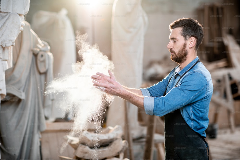 Sculpteur frappant des mains secouant la poussière après le travail dans l’atelier avec des sculptures en arrière-plan
