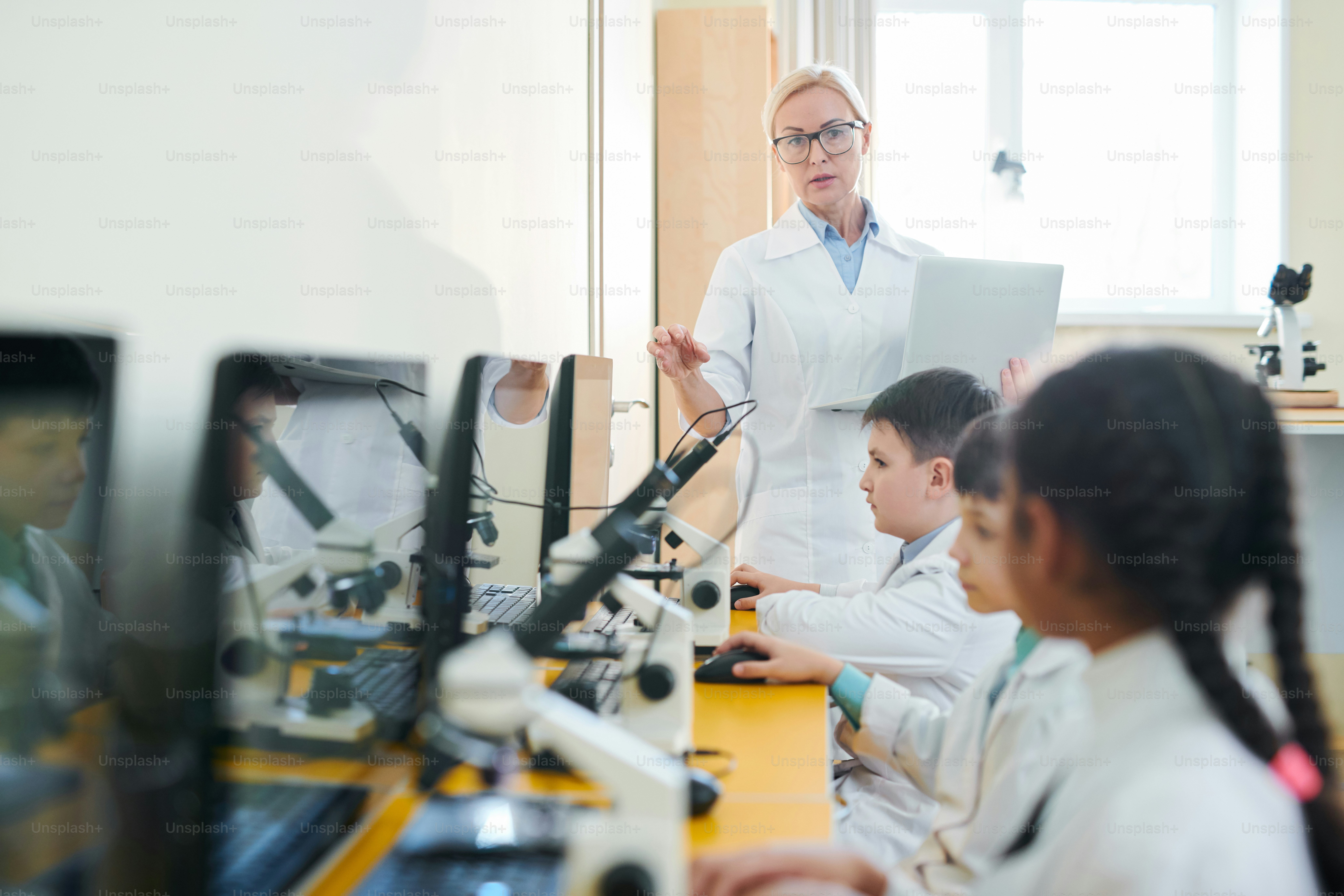Blonde mature teacher in whitecoat standing in front of her pupils at lesson and giving them instructions