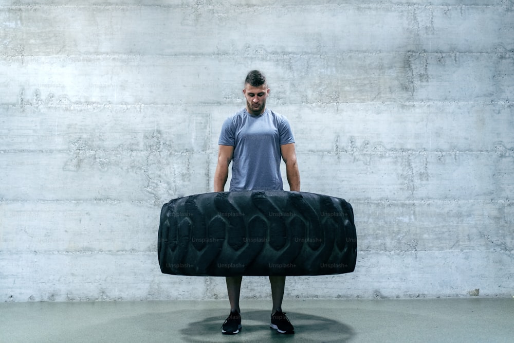 Strong muscular man in sportswear lifting tire while standing in front of the wall in gym gym.