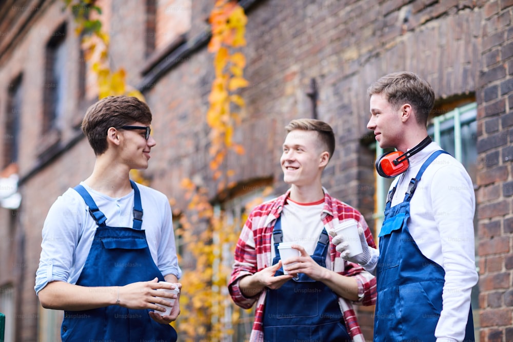 Group of positive young friends working together at factory standing in circle and drinking coffee while chatting during break outdoors