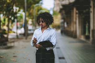 A curly Asian guy in sunglasses is belting the sleeve of his white shirt and holding a blazer from his formal suit in the hands while standing in the center of a sidewalk in Barcelona, Spain