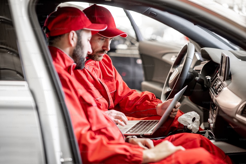 Two male auto mechanics in red uniform diagnosing car with computer sitting on the seats indoors at the car service