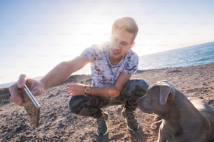 가장 친한 친구 함께 젊은 백인 남자와 회색 아름다운 amstal 개는 배경에 햇빛이 비치는 일몰이 있는 야외에서 스마트폰으로 셀카 사진을 찍고 있다 - 바다와 휴가 개념