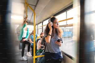 Schöne gemischtrassige Frau, die Musik hört, Smartphone benutzt und in öffentlichen Verkehrsmitteln steht.