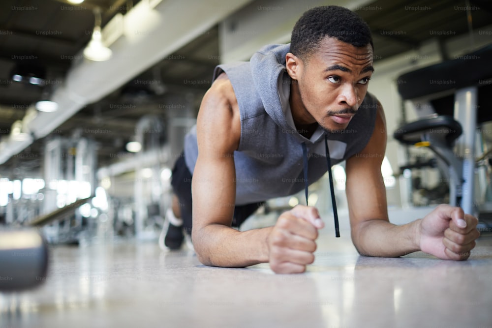 Young African-american man in activewear doing plank on the floor of training gym or fitness center