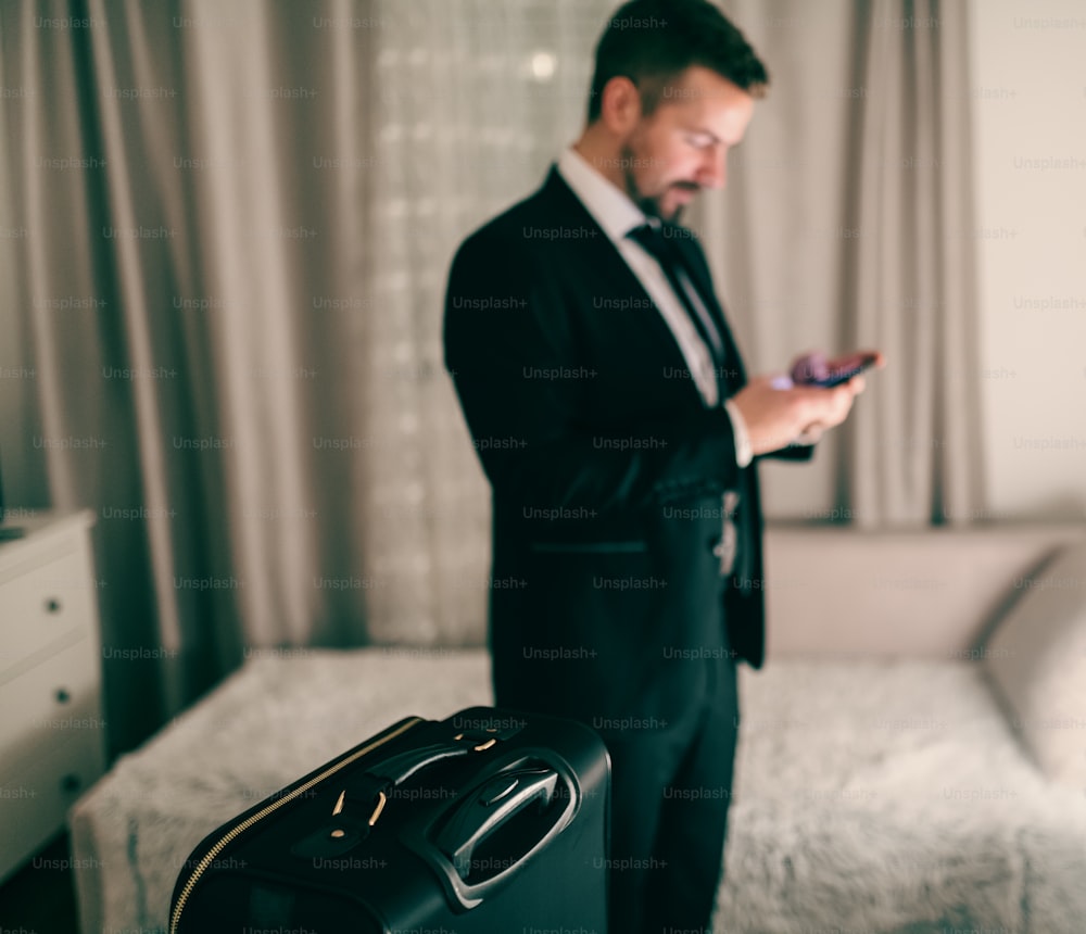 Businessman using smart phone while standing in bedroom and preparing for the trip. Selective focus on suitcase.