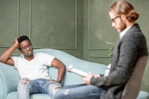 オフィスでの心理セッション中に快適なソファに座って心理学者と真剣な会話をする若いアフロ民族の男性
