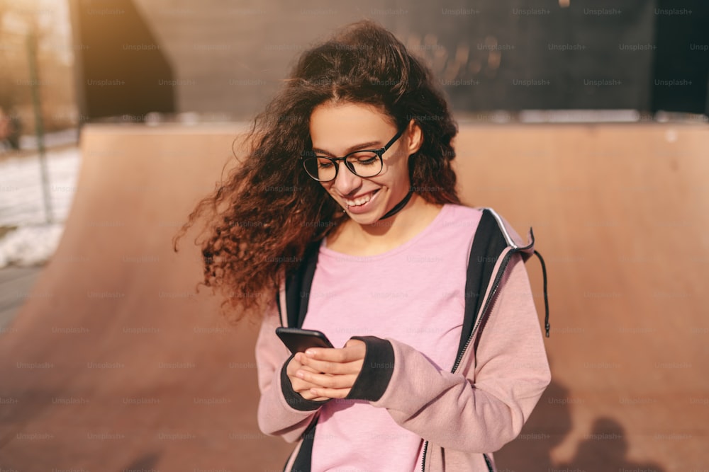Gros plan d’une jolie adolescente hipster métisse avec un sourire aux dents utilisant un téléphone intelligent tout en se tenant dans le skate park.