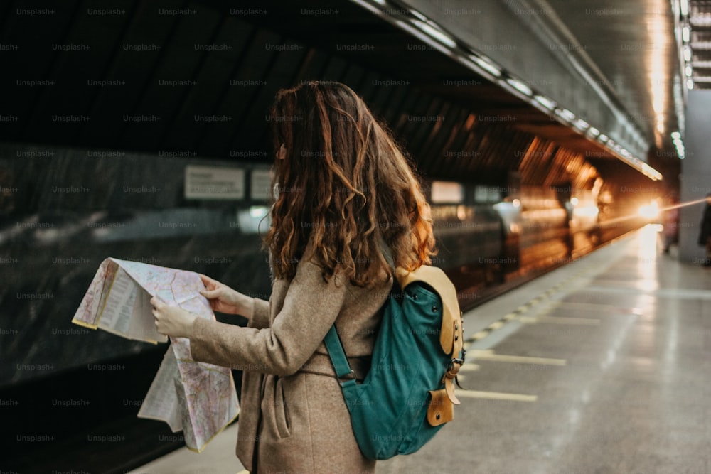 電車の前の地下鉄駅でバックパックと地図を持つ若い女性の巻き毛の赤い頭の女の子の旅行者