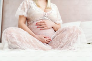 Gros plan d’une femme blonde caucasienne enceinte heureuse tenant le ventre assise avec les jambes croisées sur le lit dans la chambre.