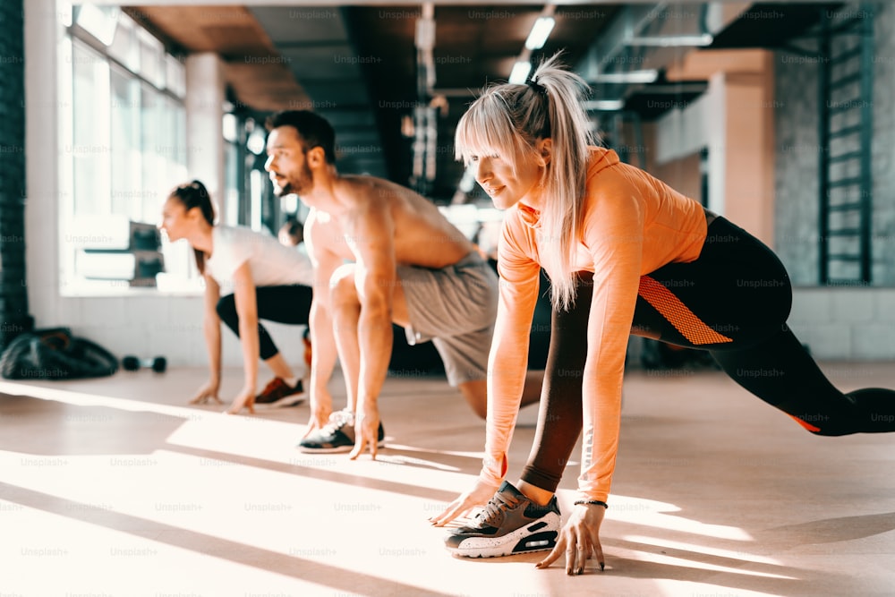 Small group of people with healthy habits doing stretching exercises on a gym floor. Selective focus on blonde woman. In background mirror.