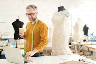 Diseñador de moda profesional maduro con cinta métrica y vaso de bebida de pie junto al escritorio frente a la computadora portátil en el estudio