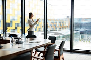 Side view portrait of elegant businesswoman looking at smartphone screen while standing by window in conference room, copy space