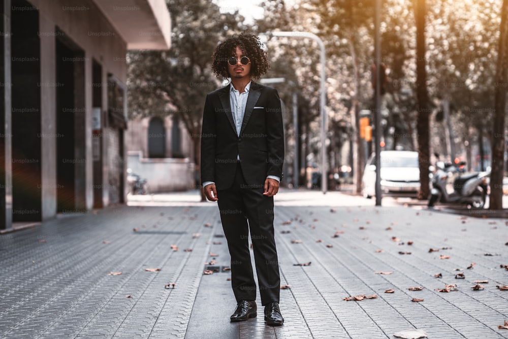 A confident Asian man entrepreneur with curly hair and in a business suit is standing in the middle of sidewalk in urban settings with buildings on the left and a road on the right