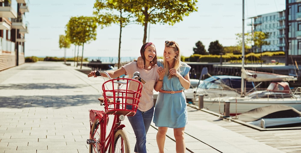Two young best friends laughing together while walking with a bicycle through the city in summer
