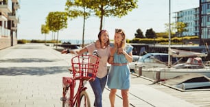 Two young best friends laughing together while walking with a bicycle through the city in summer