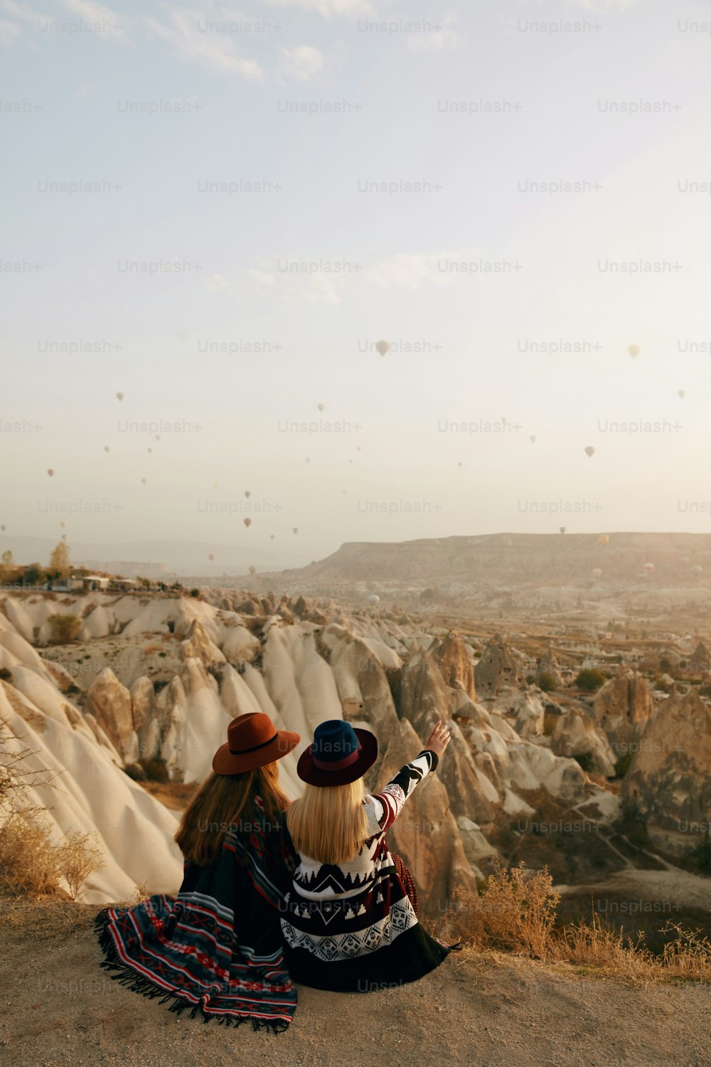 Viajar. Mujeres viajeras mirando globos aerostáticos voladores en el cielo, mujeres con sombreros sentadas en la colina disfrutando de la puesta de sol. Alta resolución