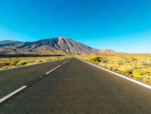 Strada lunga in montagna con monte vulcaniano di fronte e cielo limpido blu - punto di vista del suolo con asfalto nero e linee bianche - concetto di guida e viaggio