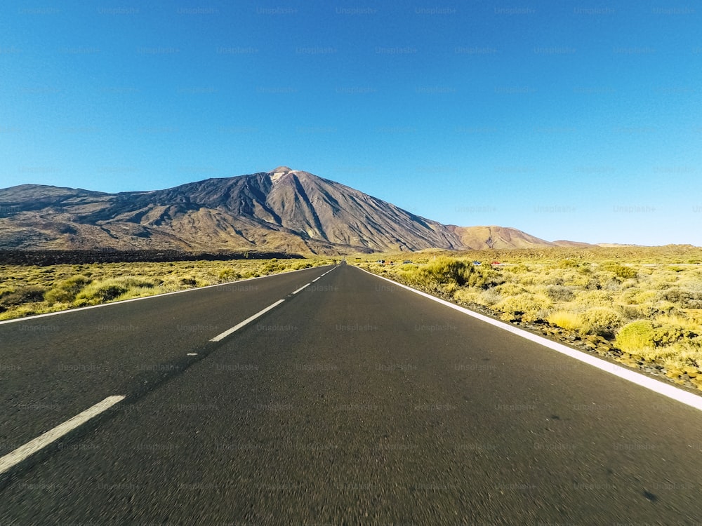 앞에 벌컨 마운트가 있고 푸른 맑은 하늘이 있는 산의 장거리 도로 - 검은색 아스팔트와 흰색 선이 있는 지상 관점 - 운전 및 여행 개념