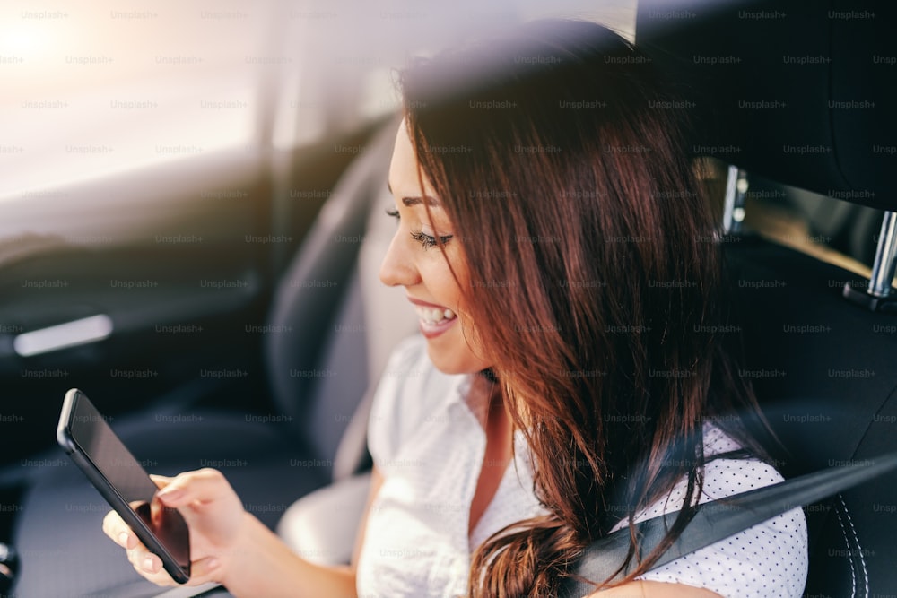Perfil de hermosa morena con sonrisa dentada usando un teléfono inteligente mientras está sentada en su automóvil.