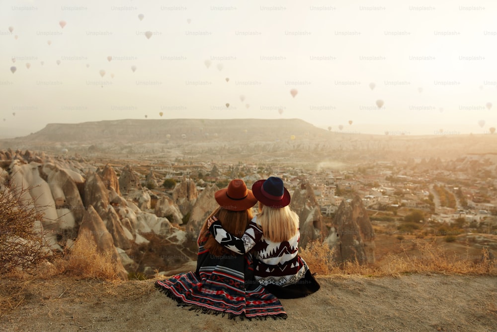 As pessoas viajam. Mulheres de chapéu sentadas na colina desfrutando de balões de ar quente voando vista na Capadócia Turquia. Alta Resolução