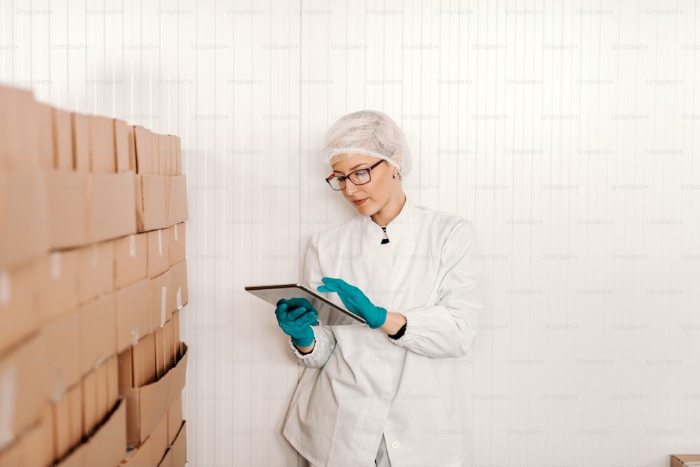 Blonde female employee in sterile uniform using tablet for logistic while standing next to boxes in food factory.