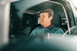 Hombre mayor caucásico serio con gorra en la cabeza sentado en un automóvil caro. Ventana abierta.